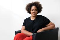 Ijeoma Oluo, an adult woman with brown skin and crown of dark, curly, ear-length hair, wearing a black shirt and orange pants, seated and looing forward