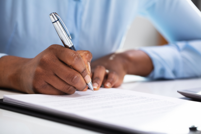 Close-up view of a person signing a contract. Person has a blue shirt and dark skin.