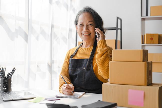 An Asian older female-presenting person talks on a cellphone while surrounded by packages package box at warehouse, merchant seller checking customer address order confirming parcel delivery